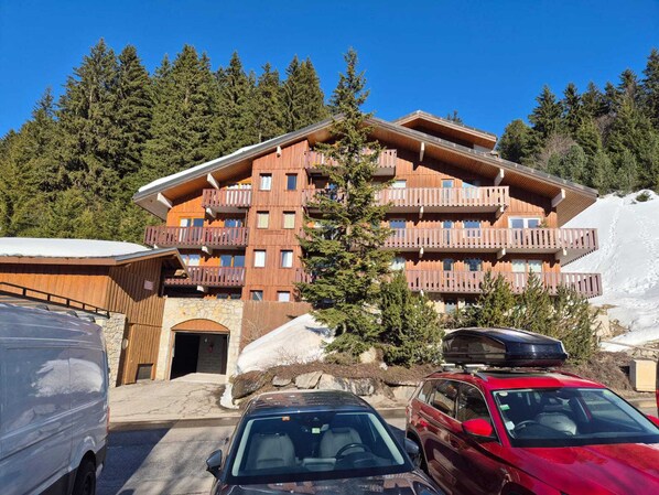 A multi-story hotel with balconies, a stone entrance, and a parking area with cars.