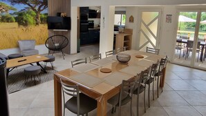 A modern dining area with a long table and chairs, a television mounted on the wall, and a view of a field through a large window. - Spacious house, private pool - 11 guests (La Flèche)