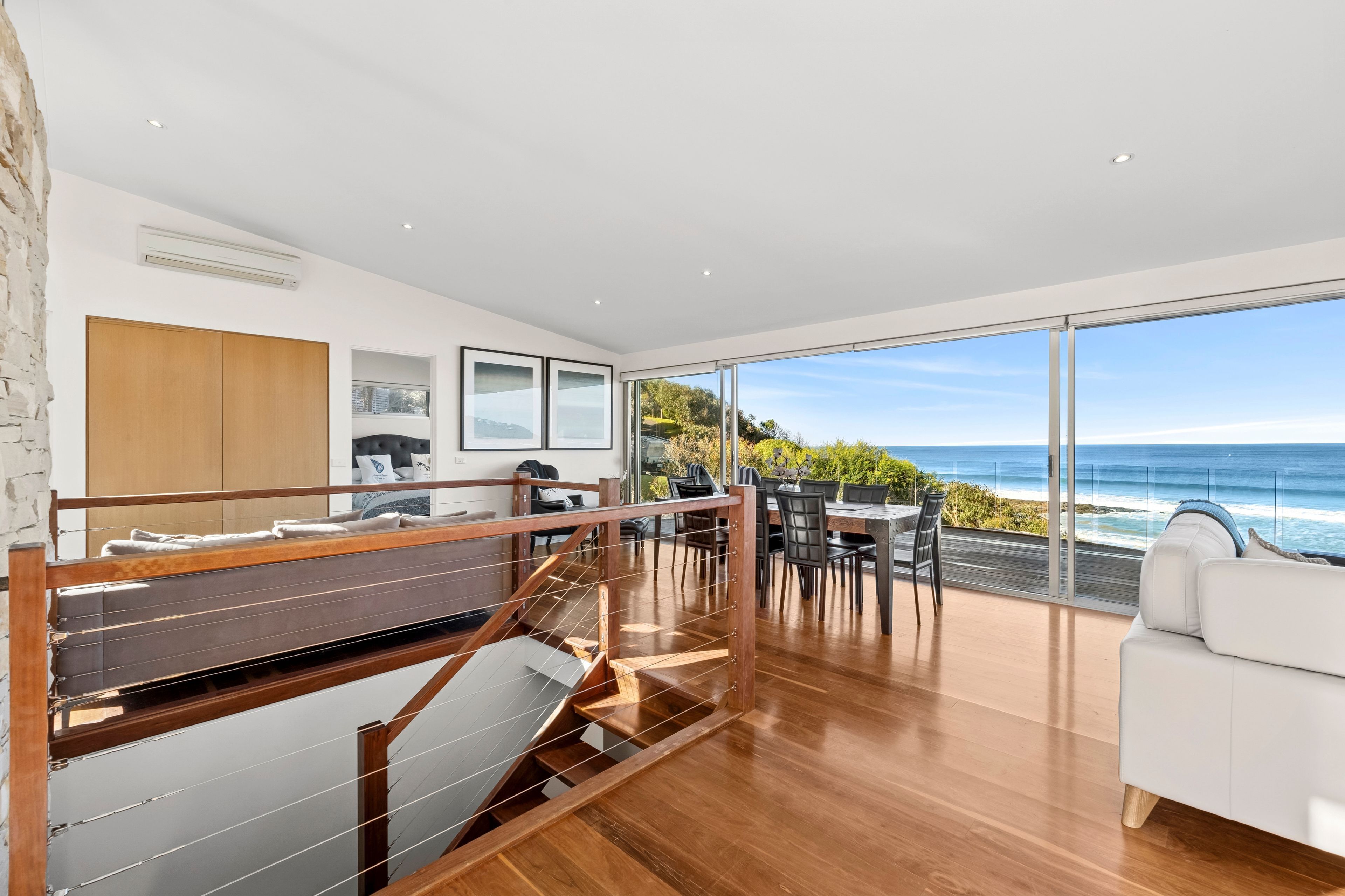 A modern interior with a wooden staircase, a dining area with a view of the ocean, and a white sofa.