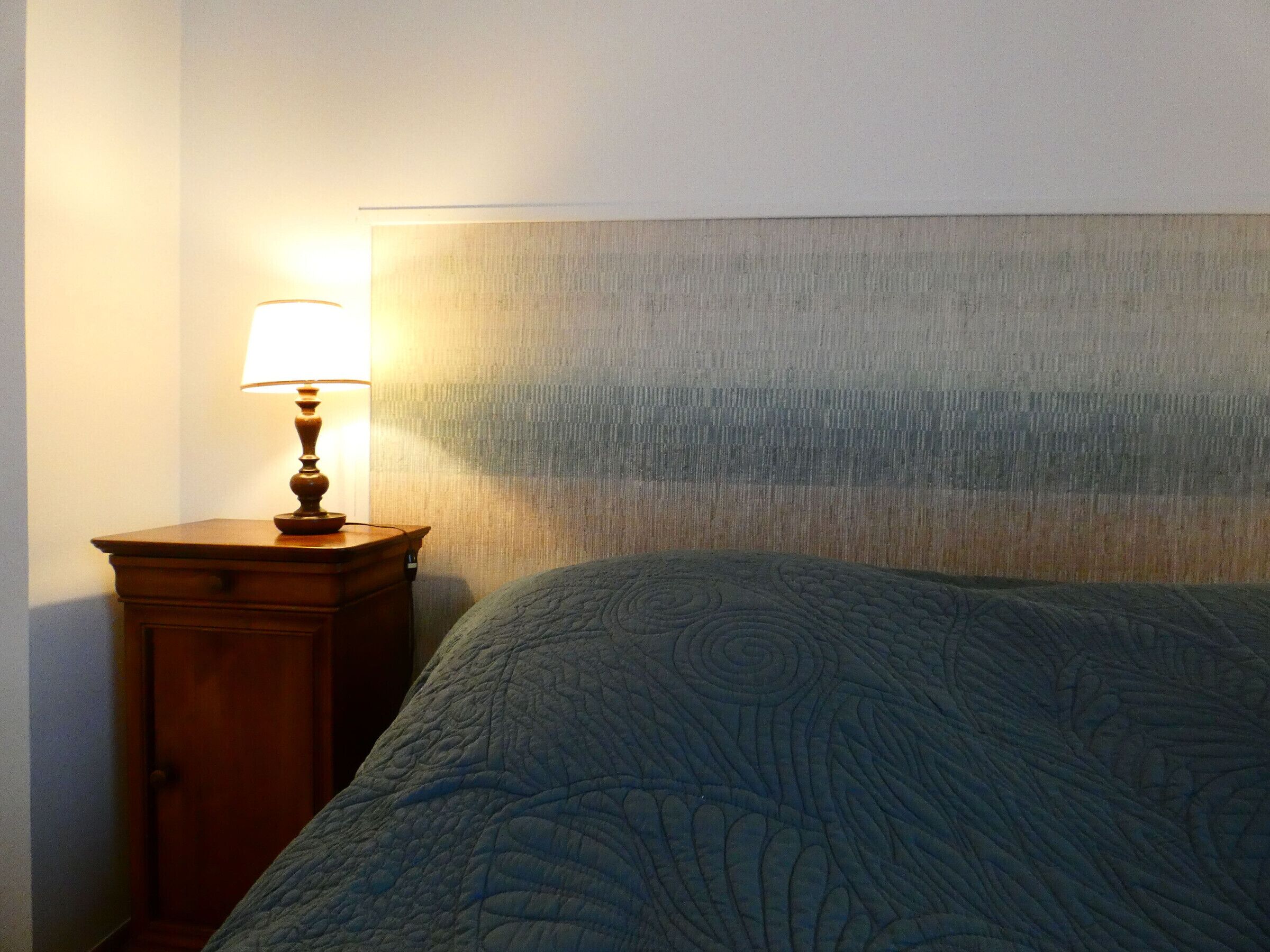 A neatly made bed with a patterned bedspread, a wooden nightstand with a lamp, and a textured headboard.