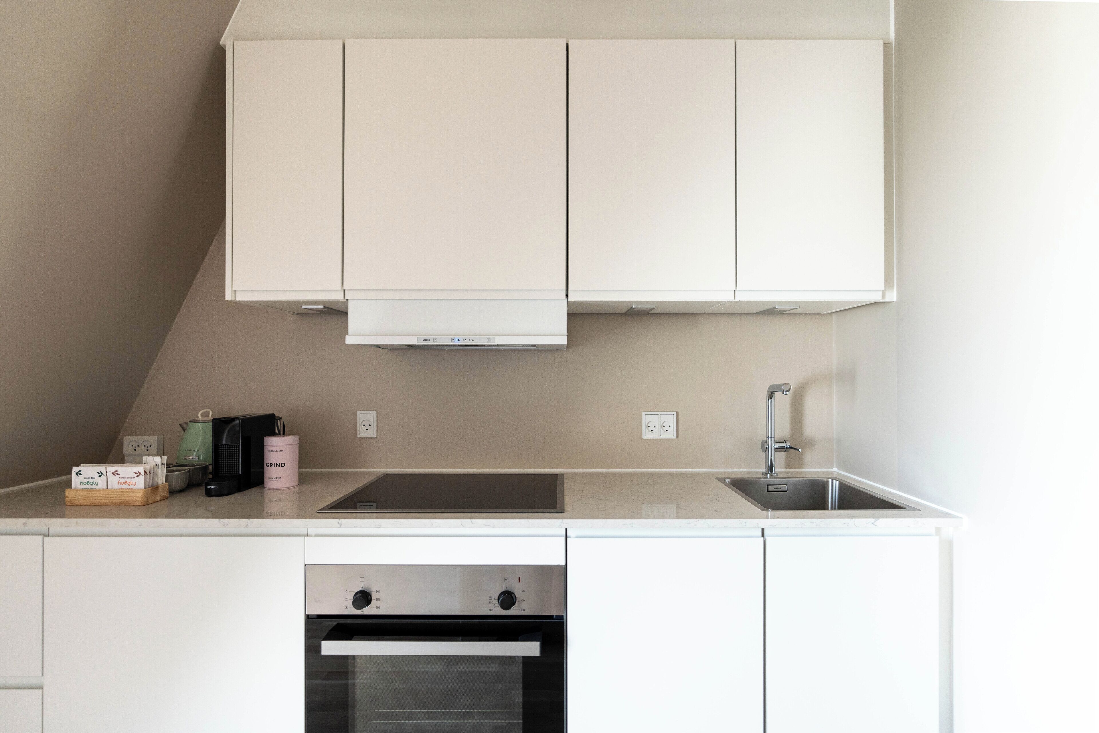 A compact kitchen with white cabinetry, a built-in oven, a sink, and a countertop with a kettle, a coffee maker, and a pink container.