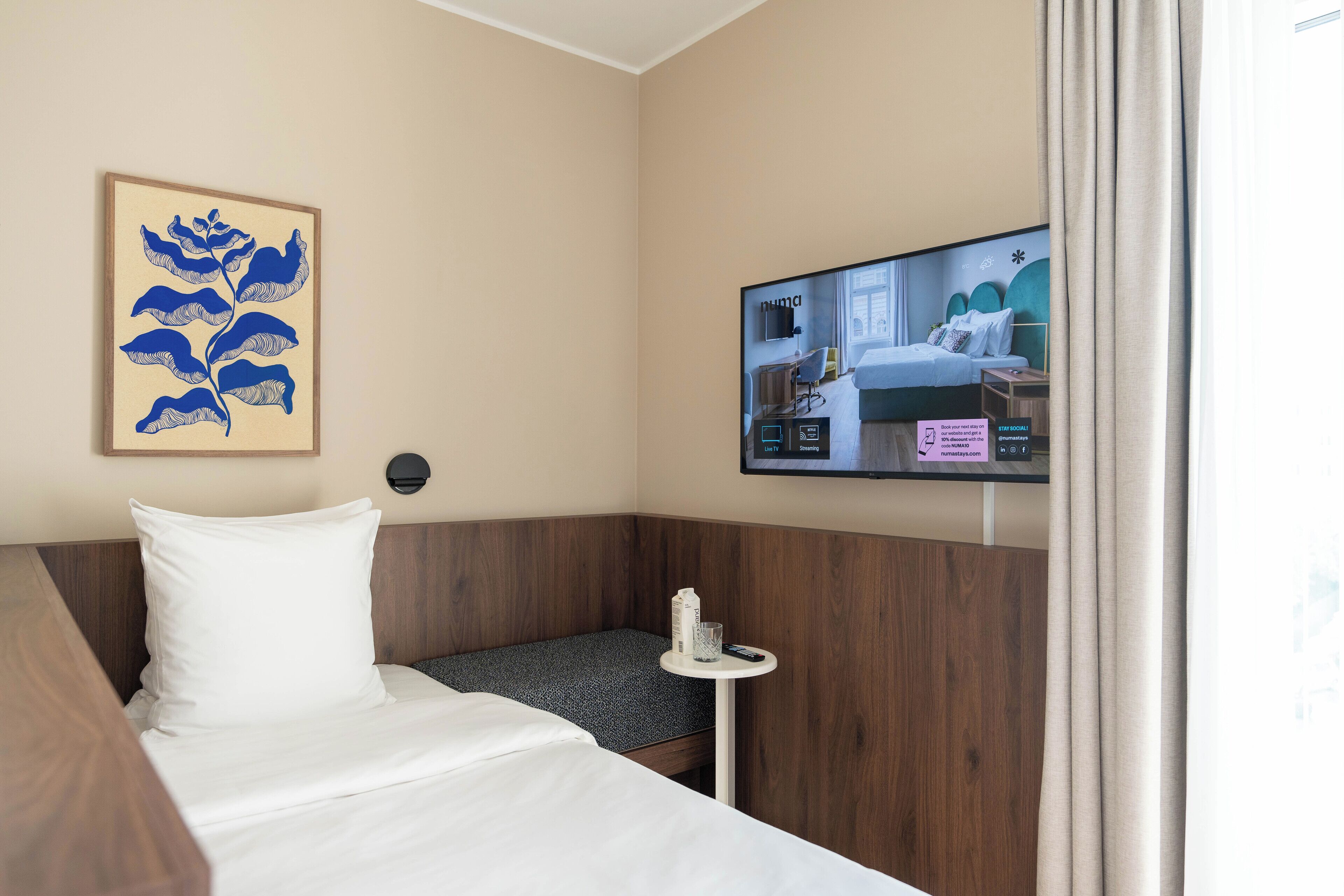 A hotel room with a bed, a small table with a glass, a pillow, a wall-mounted TV, and a framed artwork.