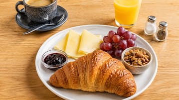 A breakfast plate with a croissant, cheese, grapes, a bowl of nuts, a glass of orange juice, and a cup of coffee on a wooden table.