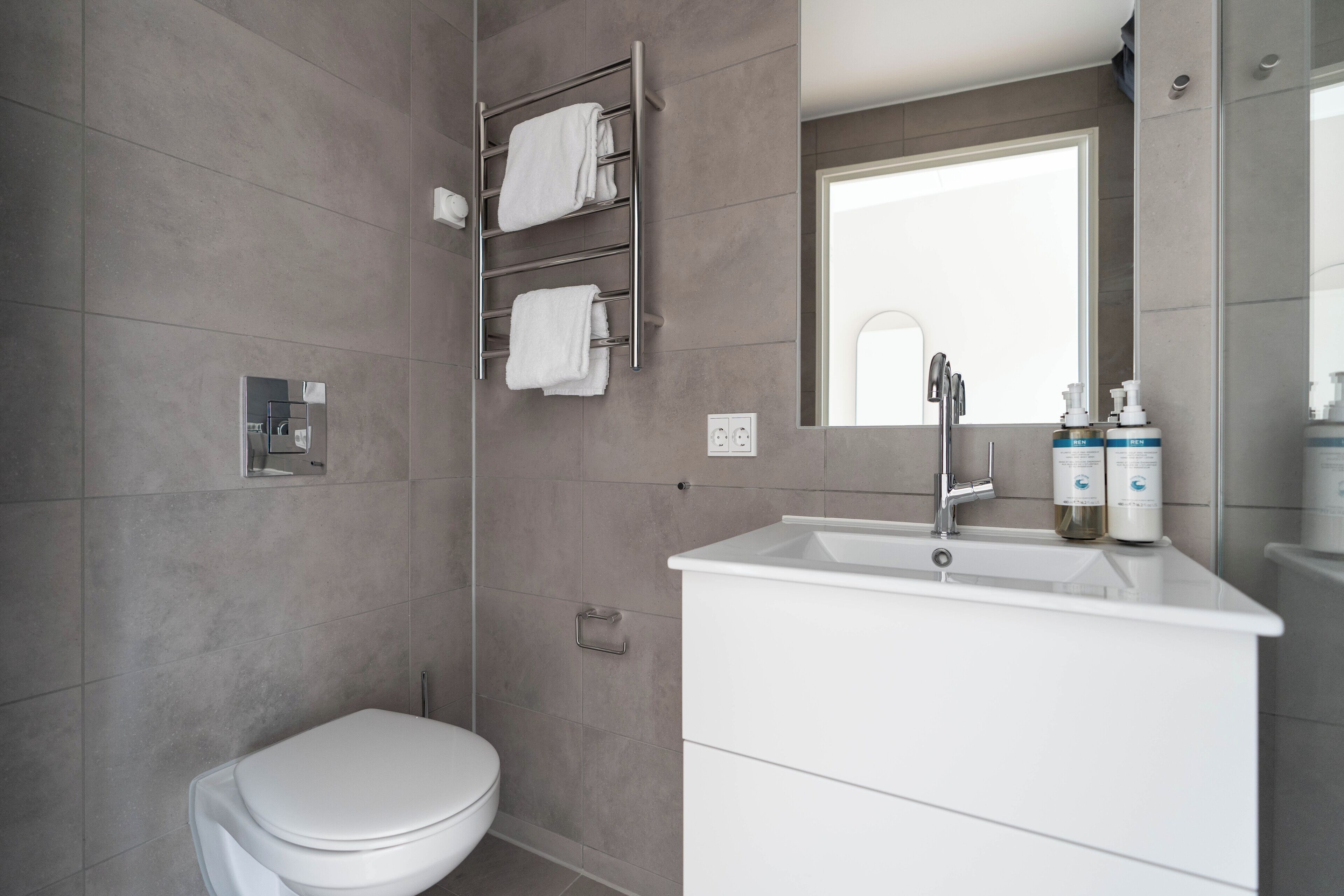Une salle de bain moderne avec des toilettes blanches, un lavabo surmonté d’un miroir et un porte-serviettes avec deux serviettes.