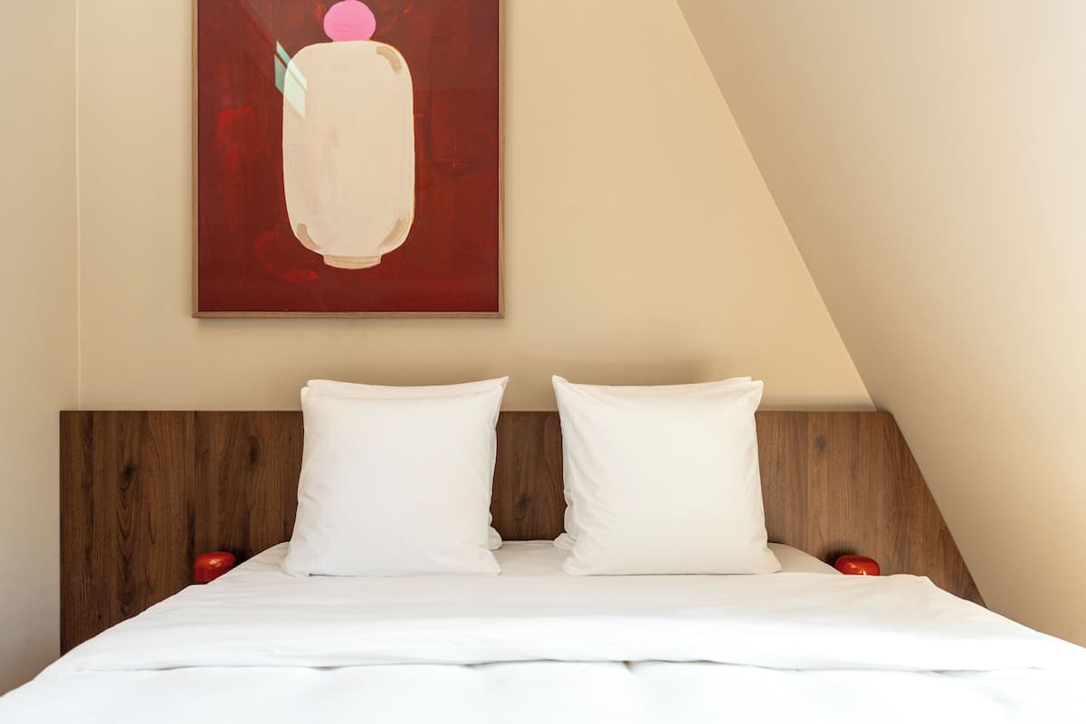 Una cama bien tendida con ropa de cama blanca y dos almohadas, un cabecero de madera y un cuadro abstracto rojo en la pared.
