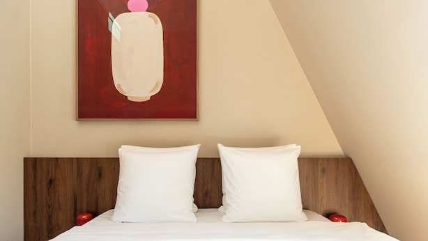 A neatly made bed with white bedding and two pillows, a wooden headboard, and a red abstract painting on the wall.