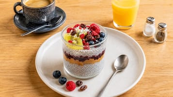 A layered chia pudding dessert with fruits and nuts on a plate, a cup of coffee, and a glass of orange juice on a wooden table.