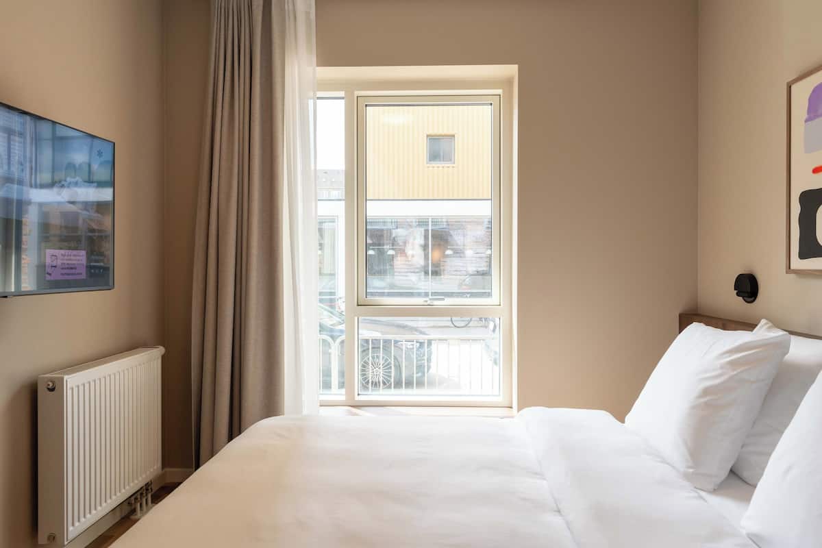 Habitación de hotel con cama, radiador, ventana con cortinas y vista al exterior.