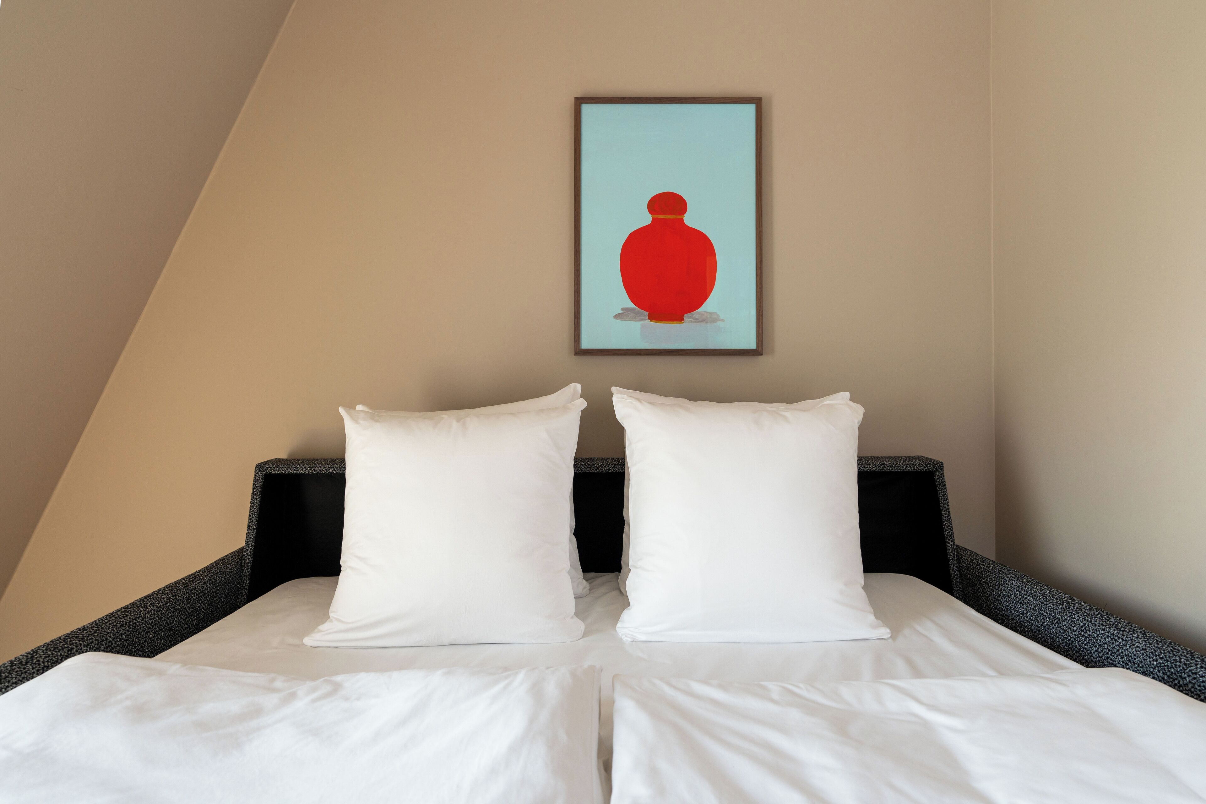 A neatly made bed with white linens and two white pillows, a framed artwork above the bed, and a dark headboard.