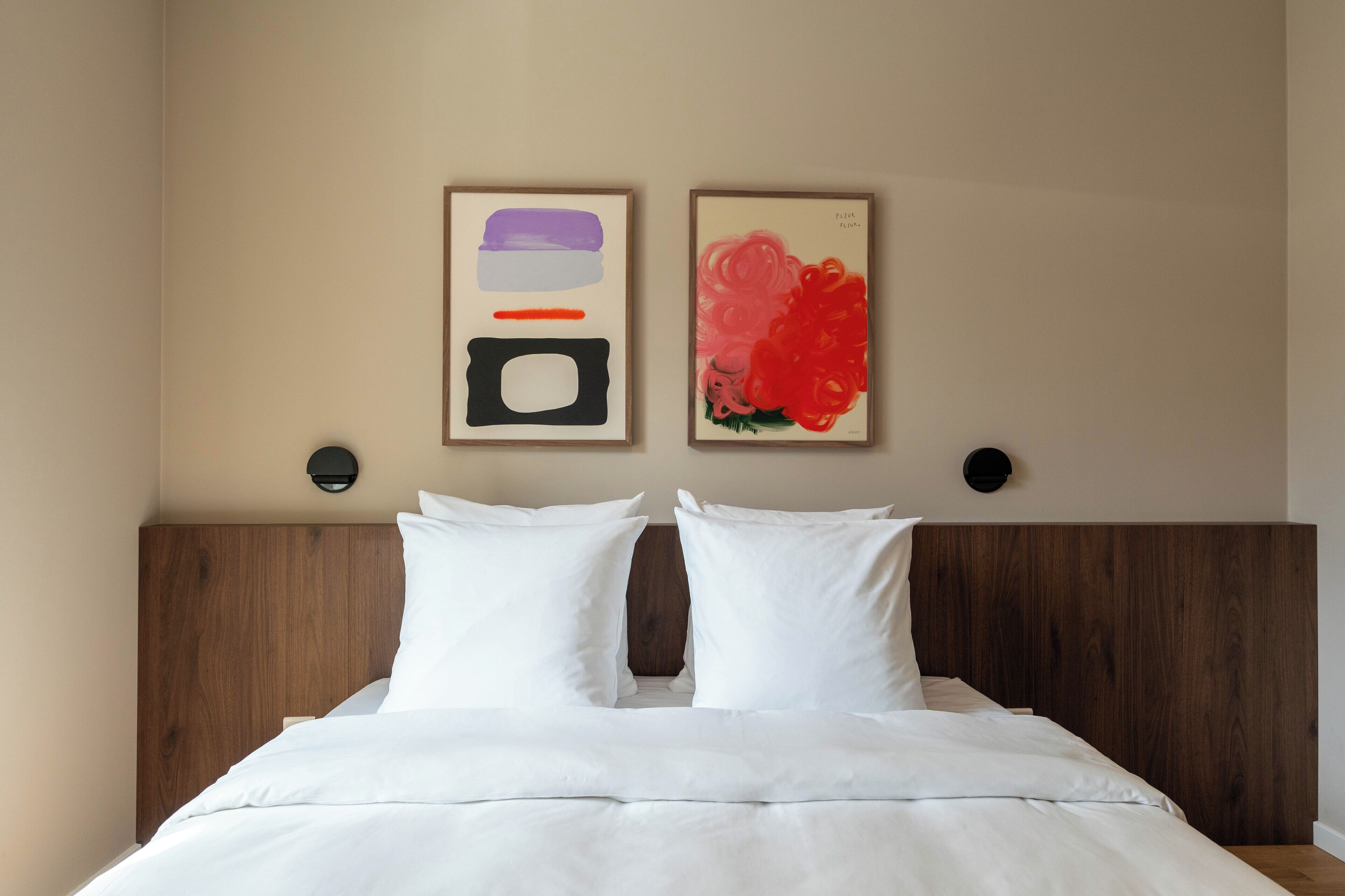 Een net opgemaakt bed met witte beddengoed en twee kussens, een houten hoofdbord en twee ingelijste kunstwerken aan de muur.