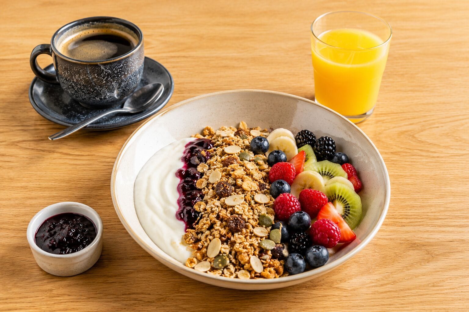 A breakfast setting with a bowl of yogurt topped with granola, berries, and sliced fruits, a cup of coffee, and a glass of orange juice on a wooden table.