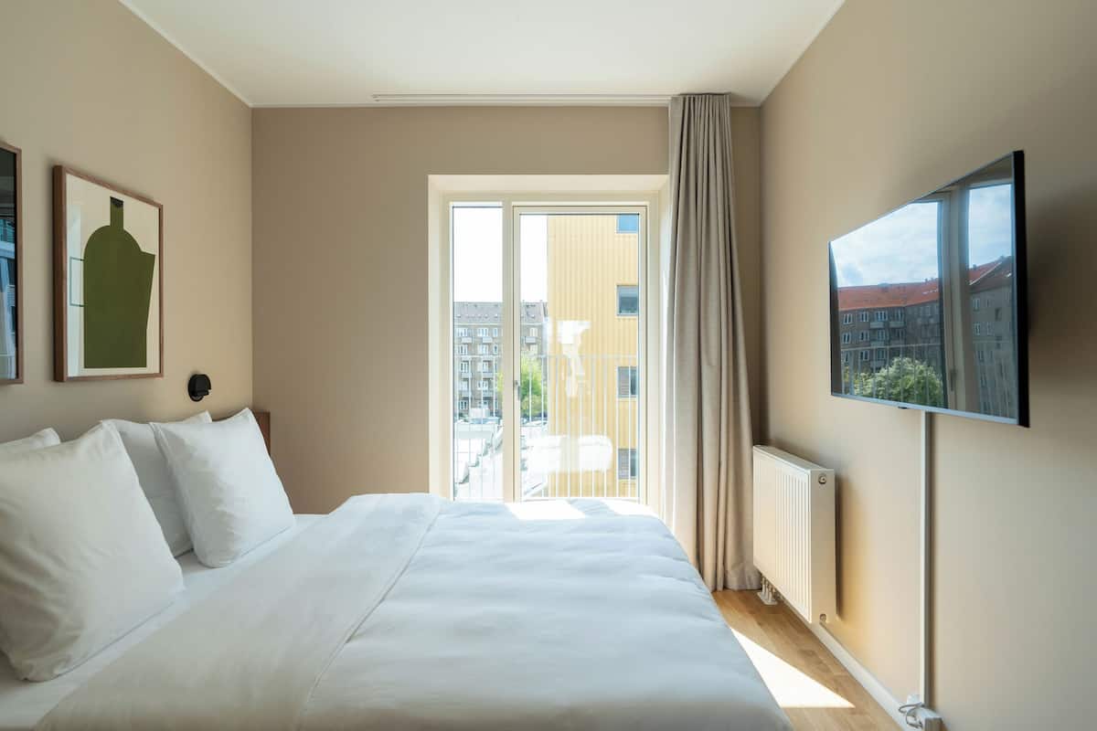 Una habitación de hotel con cama, televisor, ventana con vista a edificios y radiador.