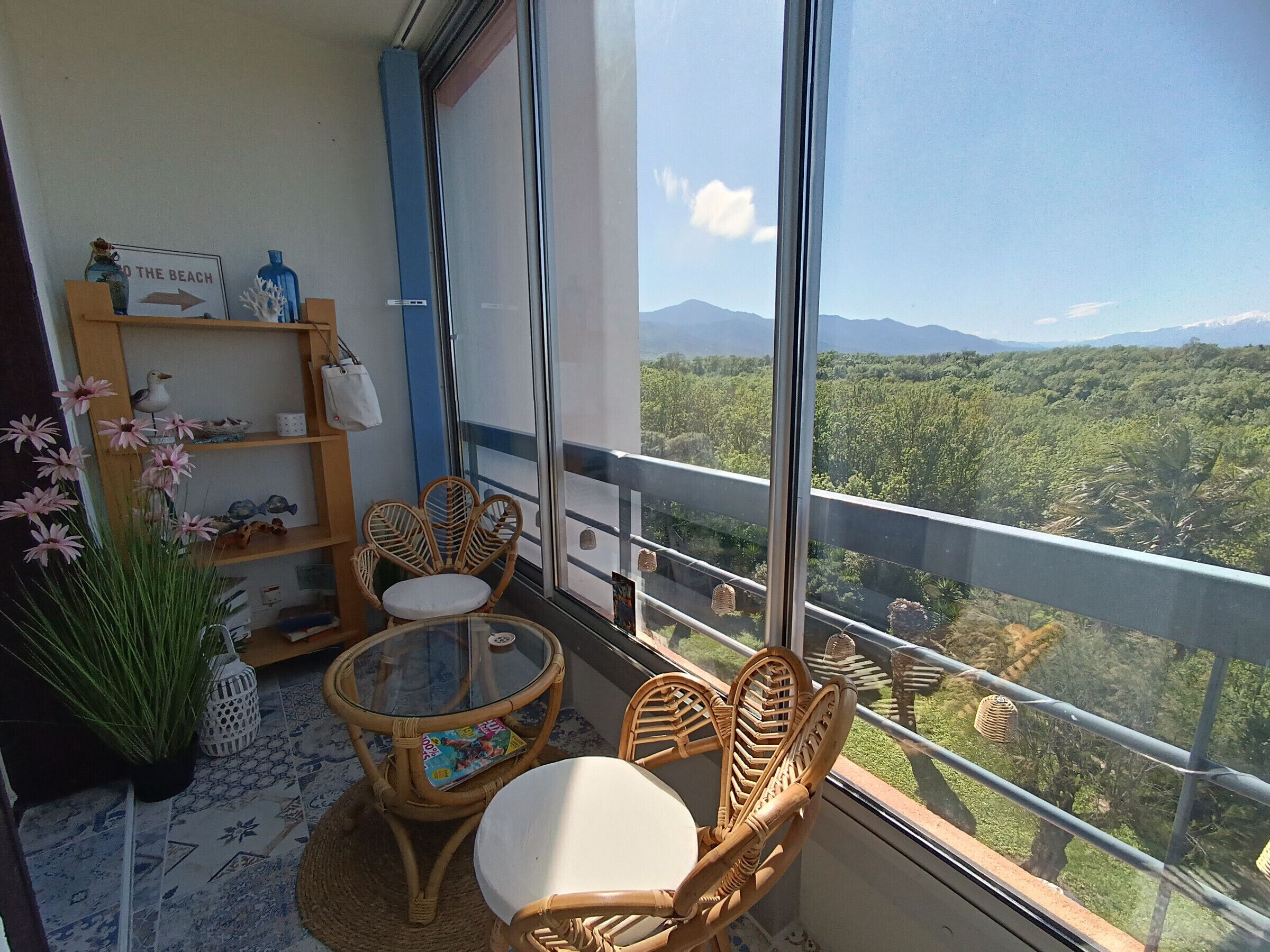 A balcony with wicker furniture, a glass table, and a view of a mountainous landscape.