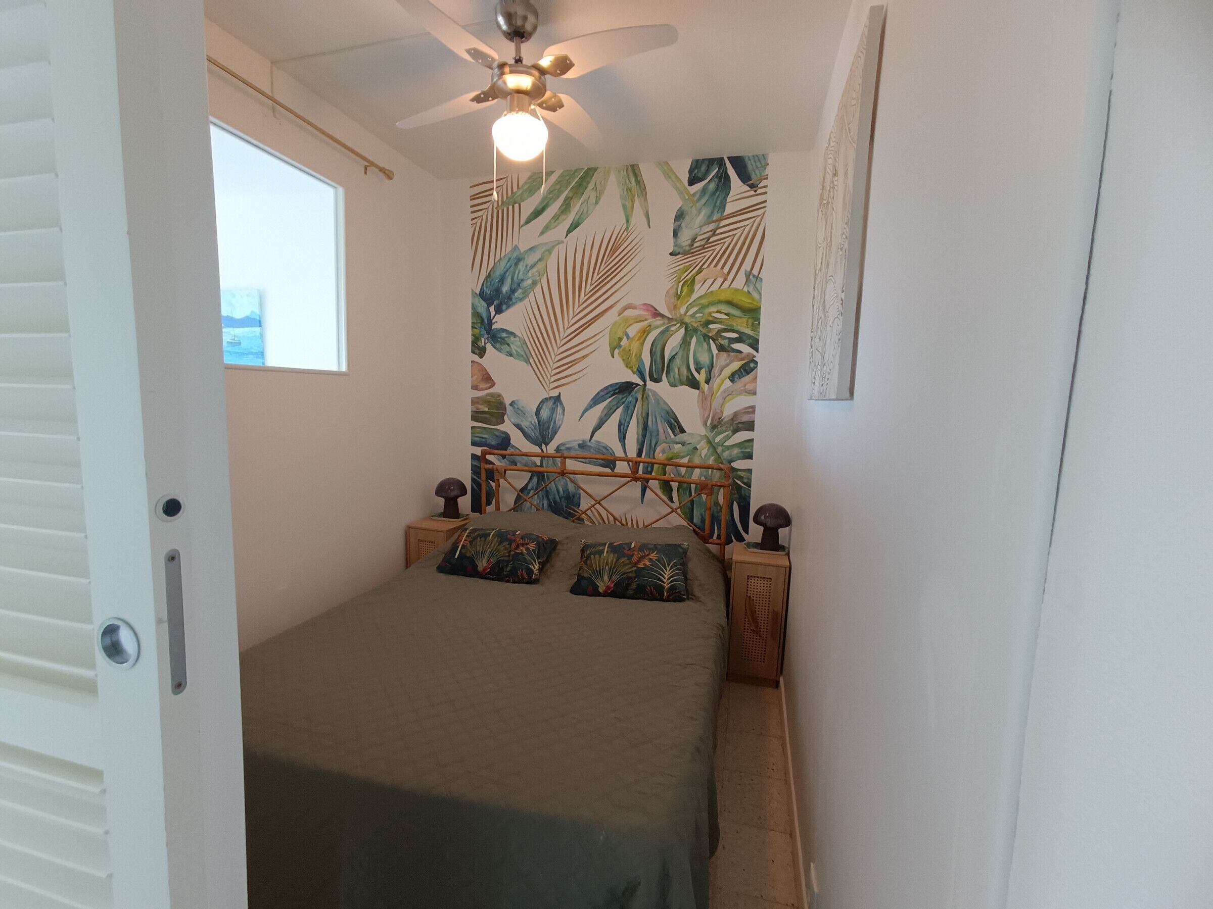 A bedroom with a bed, a ceiling fan, a window, and a tropical leaf-patterned wall.
