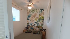 Un dormitorio con cama, ventilador de techo, ventana y pared con estampado de hojas tropicales.