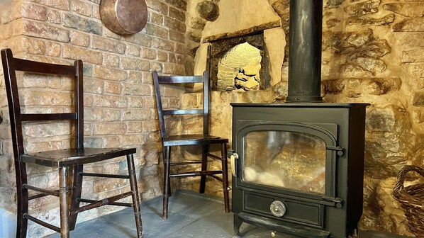A rustic room with a stone wall, a wooden chair, a wooden stool, and a black wood-burning stove.