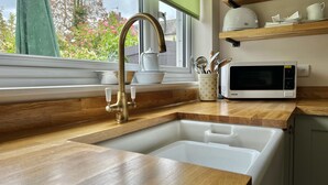Una cocina con encimera de madera, un lavamanos blanco, un grifo de latón, un microondas blanco y una ventana con vista a follaje.