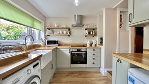 A compact kitchen with a Bosch washing machine, a microwave, and a built-in oven. There are wooden shelves above the counter and a window with a green blind. - Cozy Cottage (Wellington)