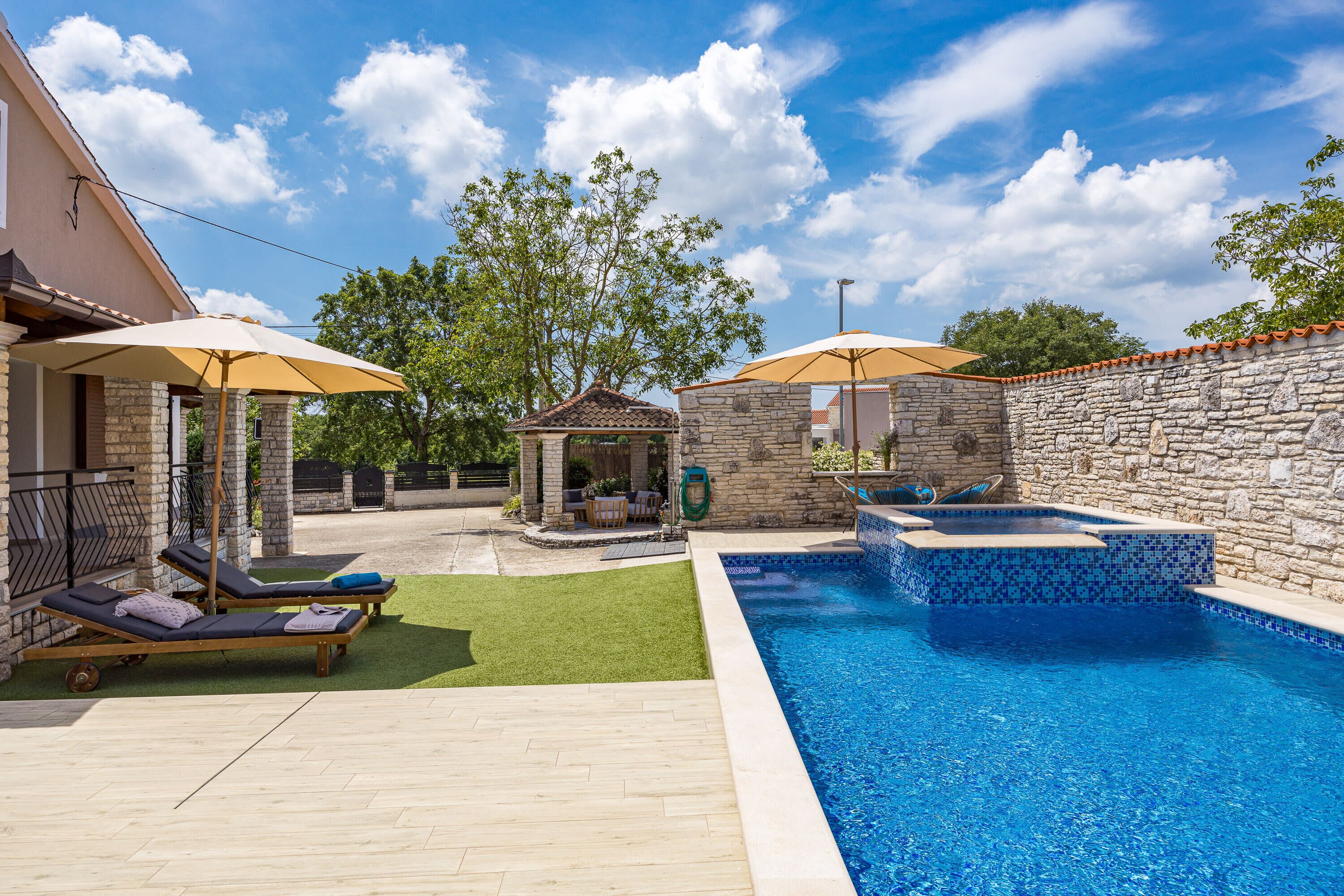 A swimming pool with a stone wall and a gazebo, surrounded by a patio with lounge chairs and umbrellas.