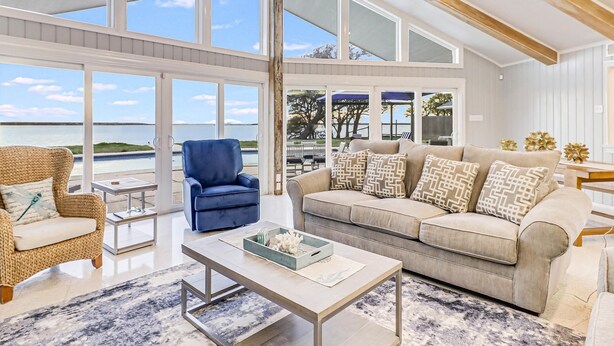 Ein geräumiges Wohnzimmer mit einem großen Fenster mit Blick auf ein Gewässer, ein blauer Sessel, ein beiges Sofa mit gemusterten Kissen und ein Couchtisch.
