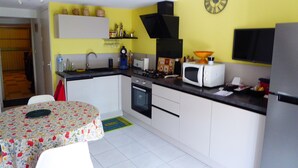 A compact kitchen with white cabinets, a microwave, a stove, and a sink. There's a clock on the wall and a small table with a floral tablecloth. - Air-Conditioned Cottage with Large Terrace, Near Beach and Shops (Argelès-sur-Mer)
