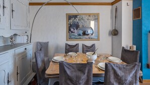 Ein Essbereich mit einem Holztisch, gedeckt für vier Personen, umgeben von grauen Polsterstühlen. Eine weiße Kommode mit aufwendigen Verzierungen befindet sich links, und eine blaue Akzentwand rechts.