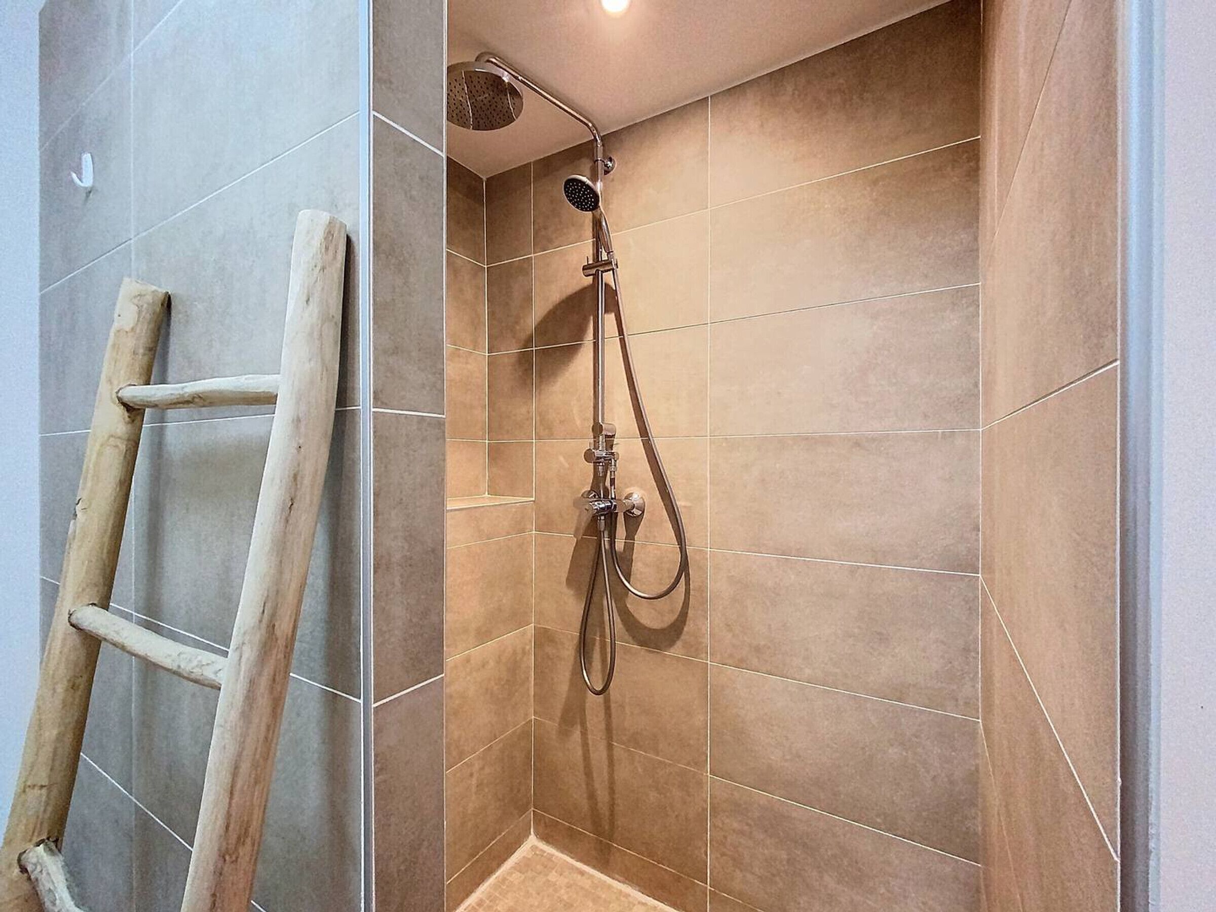 Zona de ducha con azulejos color beige, una escalera de madera y un cabezal de ducha de mano.
