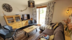 Ein gemütliches Wohnzimmer mit Sofa, Sesseln, einem Fernseher, Holzschränken und einer großen Wanduhr.
