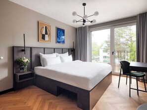 A modern hotel room with a large bed, a bedside table with a vase of flowers, a desk with a chair, and a balcony with a view of trees. - Numa | Munich Obersendling (Munich)