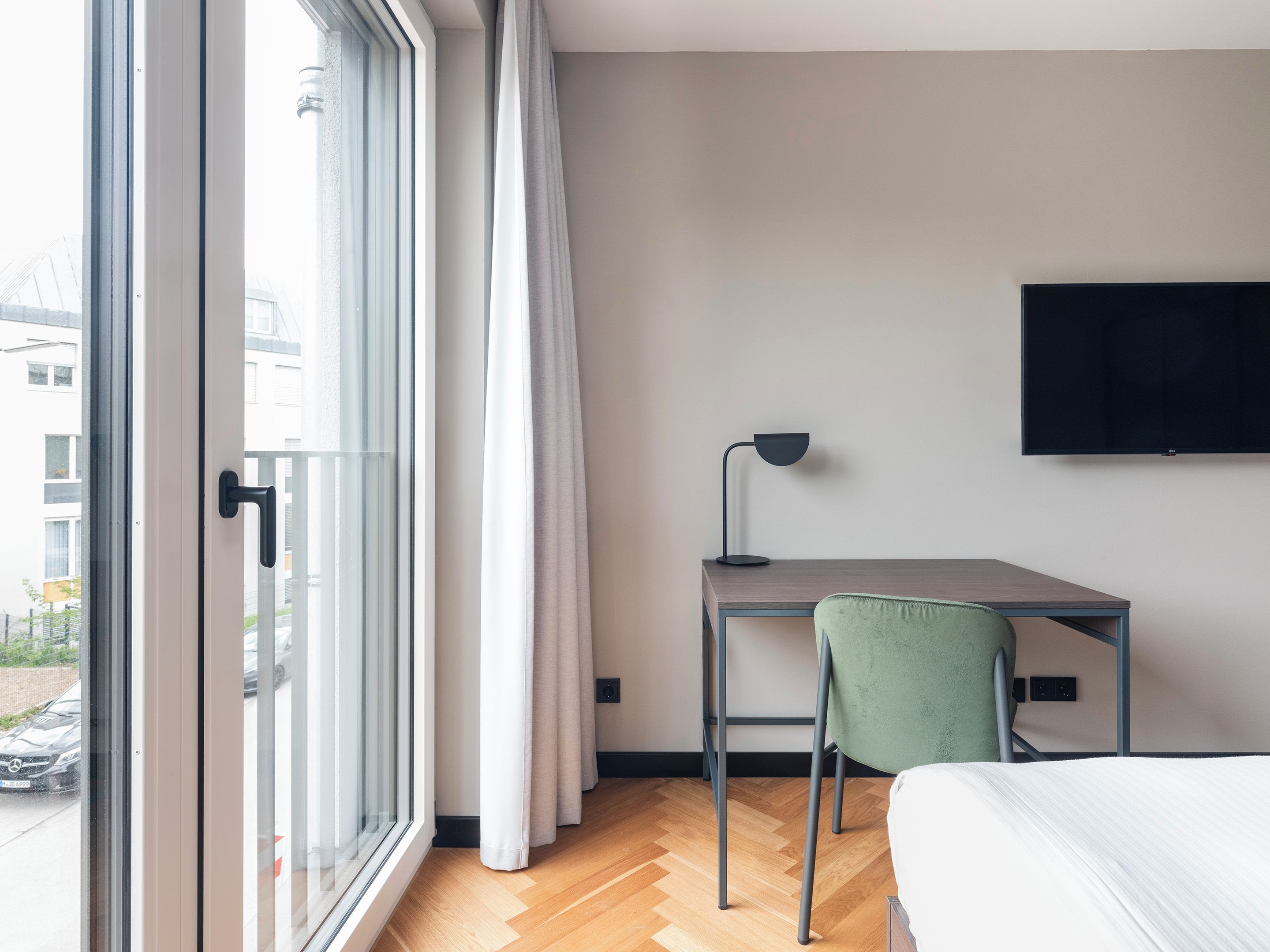 A modern hotel room with a large window, a desk with a lamp, a green chair, and a bed.