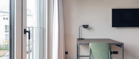 A modern hotel room with a large window, a desk with a lamp, a green chair, and a bed.