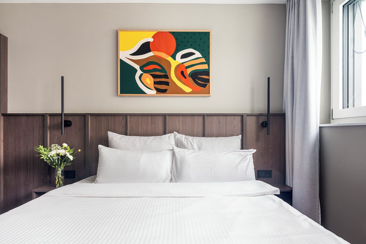 A modern hotel room with a large bed, a wooden headboard, a wall-mounted abstract painting, and a window with curtains.