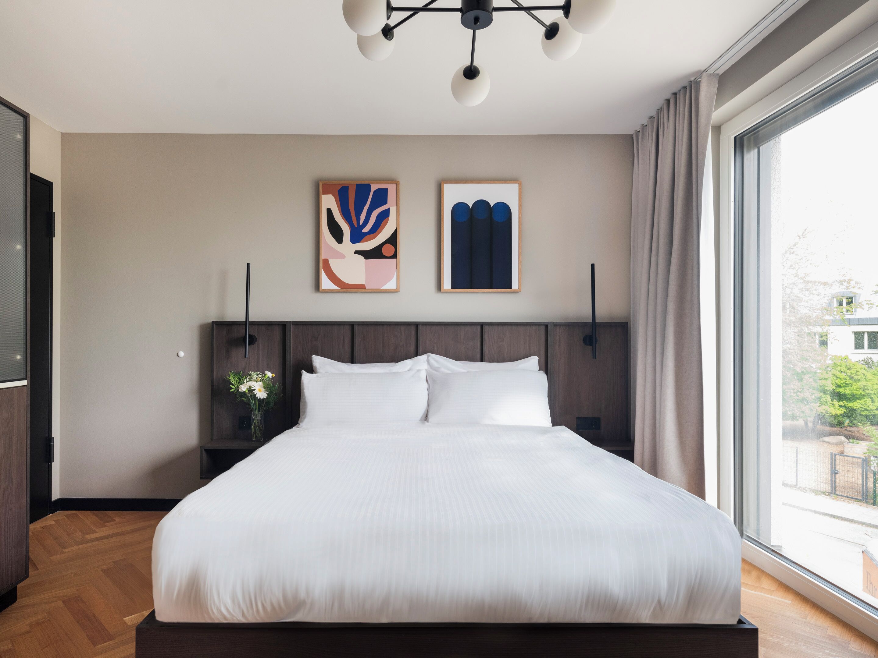 Ein modernes Hotelzimmer mit einem großen Bett, einem Holzkopfende, zwei abstrakten Bildern, einem Nachttisch mit Blumen in einer Vase und einem großen Fenster mit Blick ins Freie.