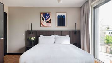 Ein modernes Hotelzimmer mit einem großen Bett, einem Holzkopfende, zwei abstrakten Bildern, einem Nachttisch mit Blumen in einer Vase und einem großen Fenster mit Blick ins Freie.