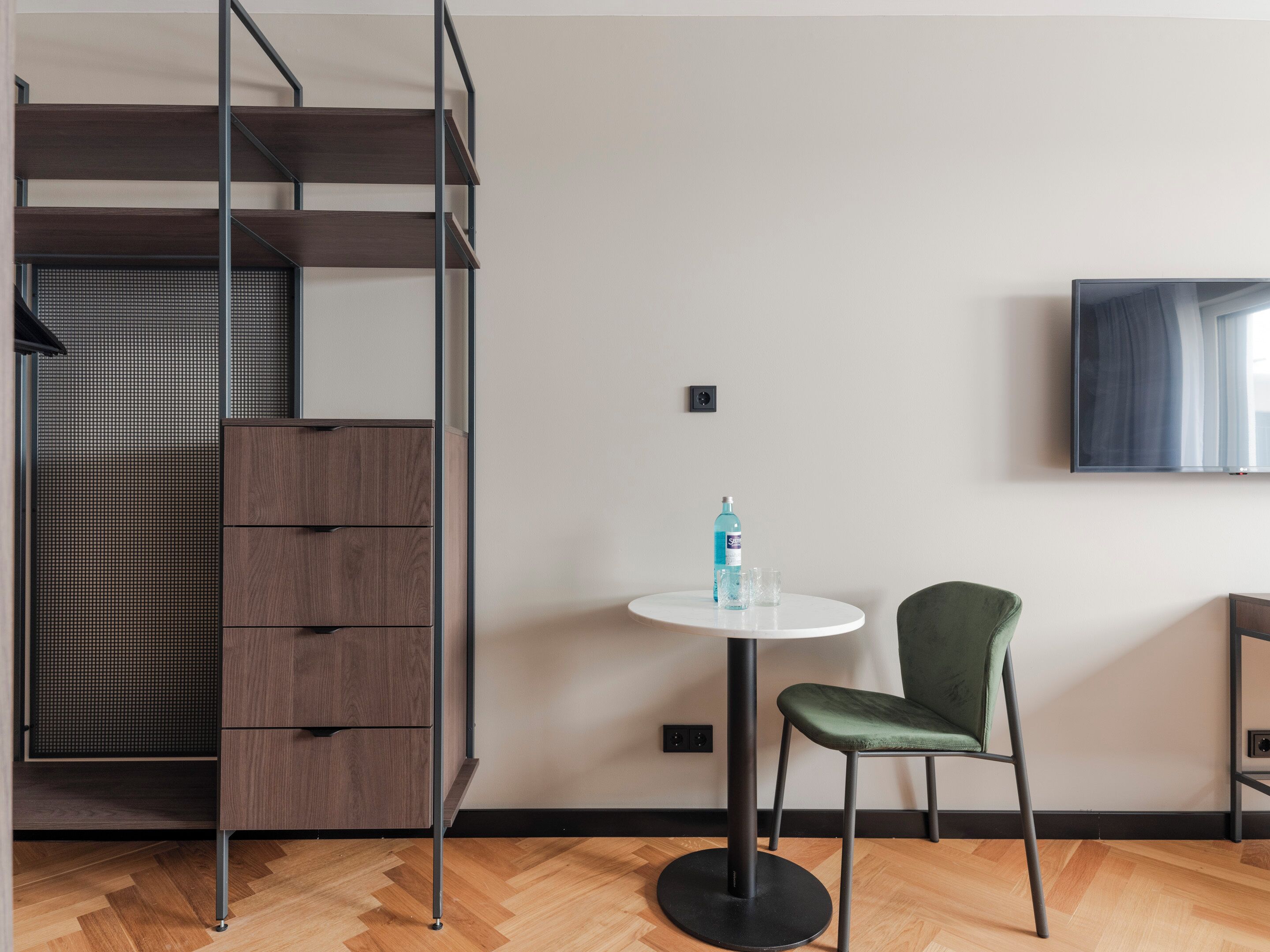 Una habitación moderna con una cama litera, una mesita redonda con una botella y vasos, una silla verde y un televisor en la pared.