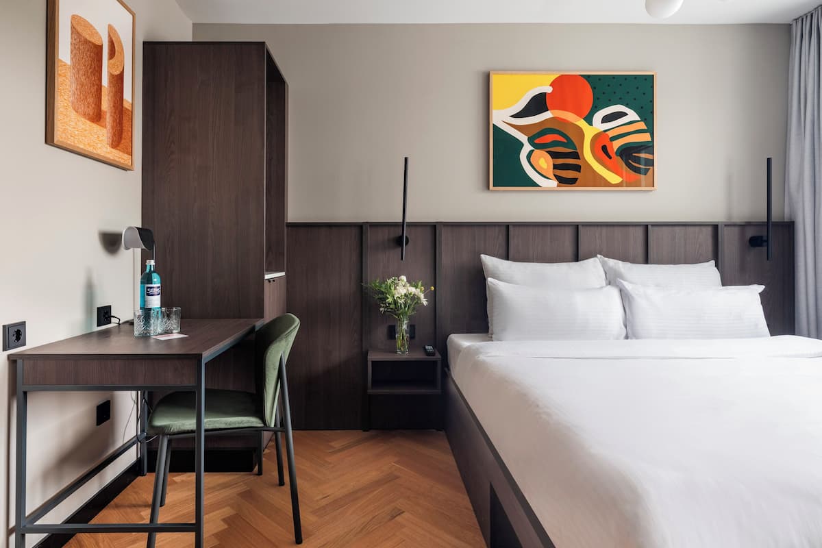 A hotel room with a bed, desk, chair, nightstand, and artwork on the wall.