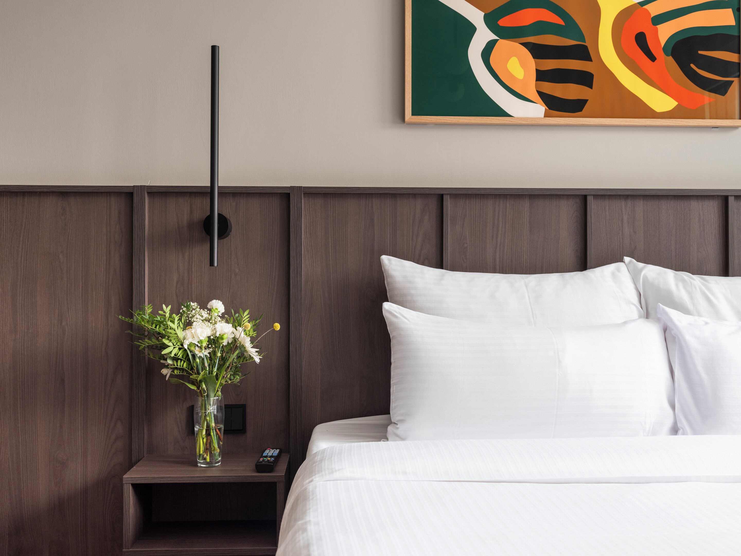A hotel room with a wooden headboard, a bedside table with a vase of flowers, and a remote control.