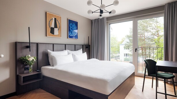 A modern hotel room with a large bed, a bedside table with a vase of flowers, a desk with a chair, and a balcony with a view of trees.