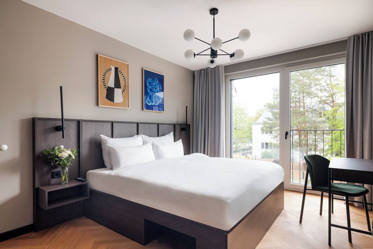 A modern hotel room with a large bed, a bedside table with a vase of flowers, a desk with a chair, and a balcony with a view of trees.