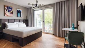 A modern hotel room with a large bed, a small table with a chair, a TV, and a balcony with curtains.