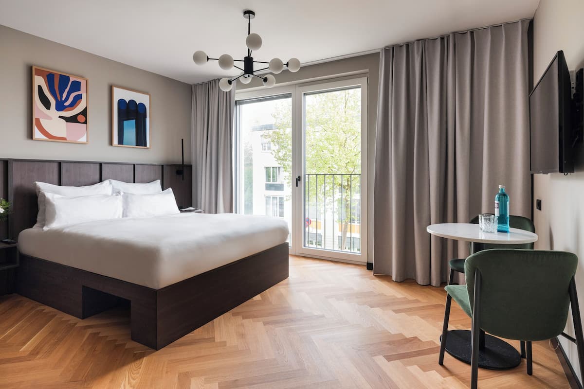 A modern hotel room with a large bed, a small table with a chair, a TV, and a balcony with curtains.