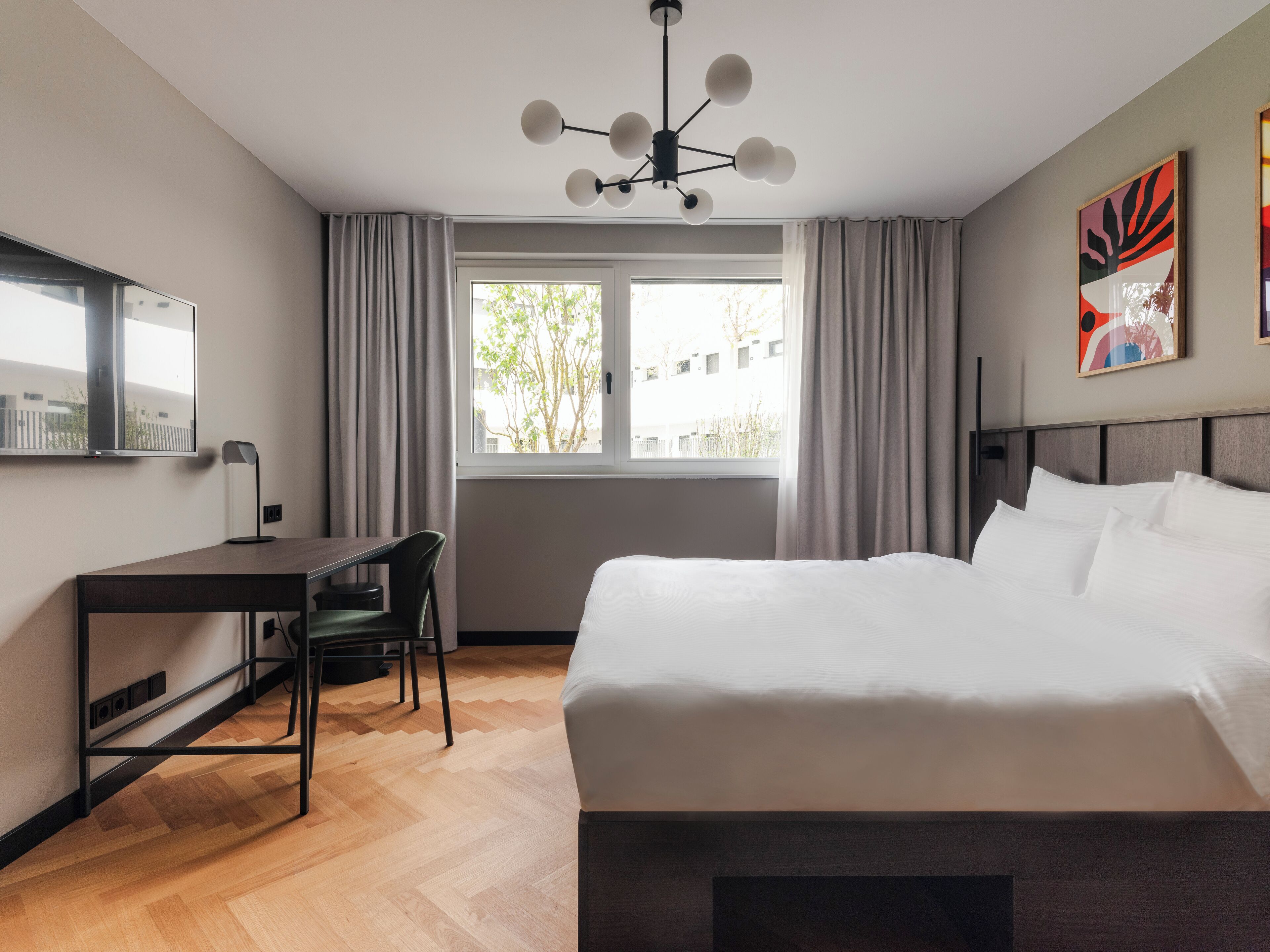 Ein modernes Hotelzimmer mit einem großen Bett, einem Schreibtisch mit Stuhl, einer Deckenleuchte und einem Fenster mit Vorhängen.