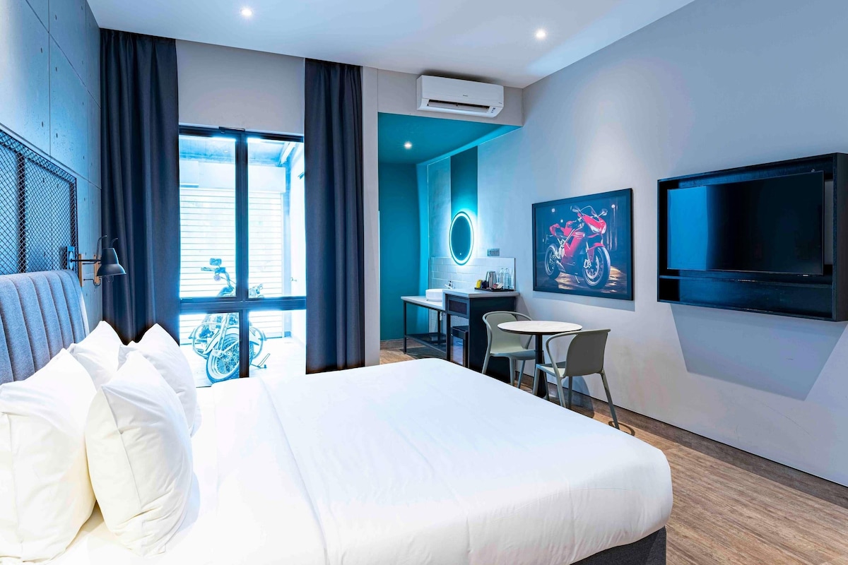 A modern hotel room with a large bed, a desk, a chair, a TV, and an air conditioning unit.