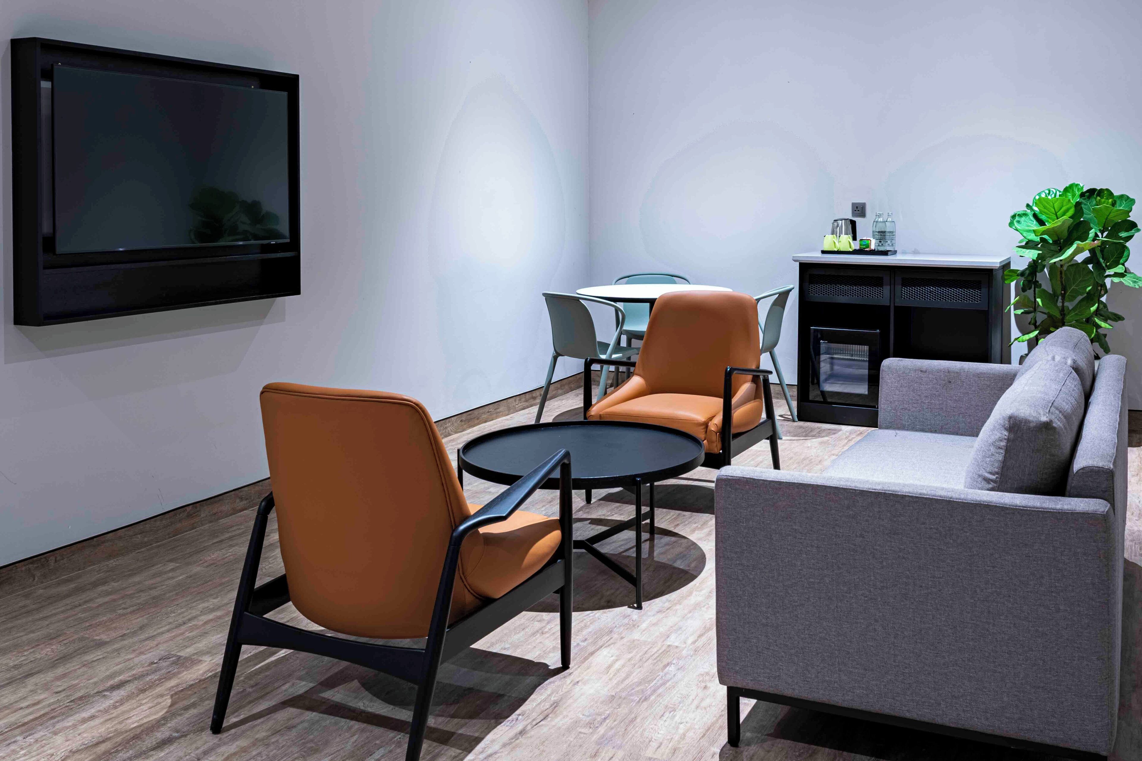 Een moderne woonkamer met een flatscreen televisie aan de muur, twee oranje fauteuils, een grijze bank en een kleine ronde tafel.