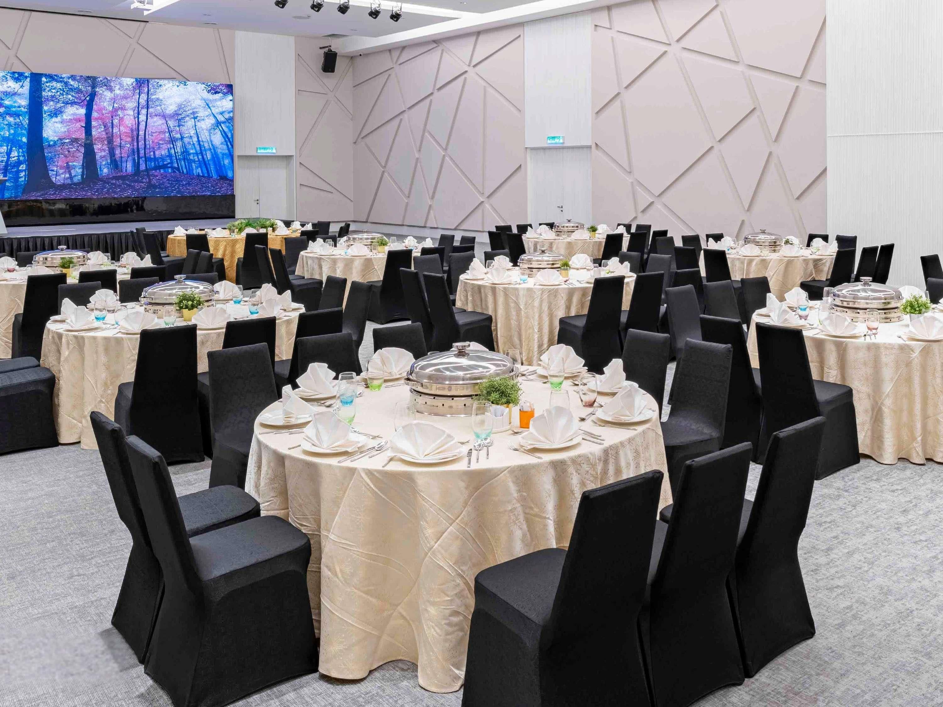 Ein Festsaal mit runden Tischen, schwarzen Stühlen und einer großen Leinwand, die eine Waldszene zeigt.