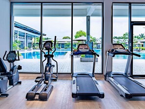 A gym with treadmills, an elliptical, and a stationary bike, overlooking a swimming pool and a building with a modern design. - Ibis Styles Sepang Klia (Sepang)