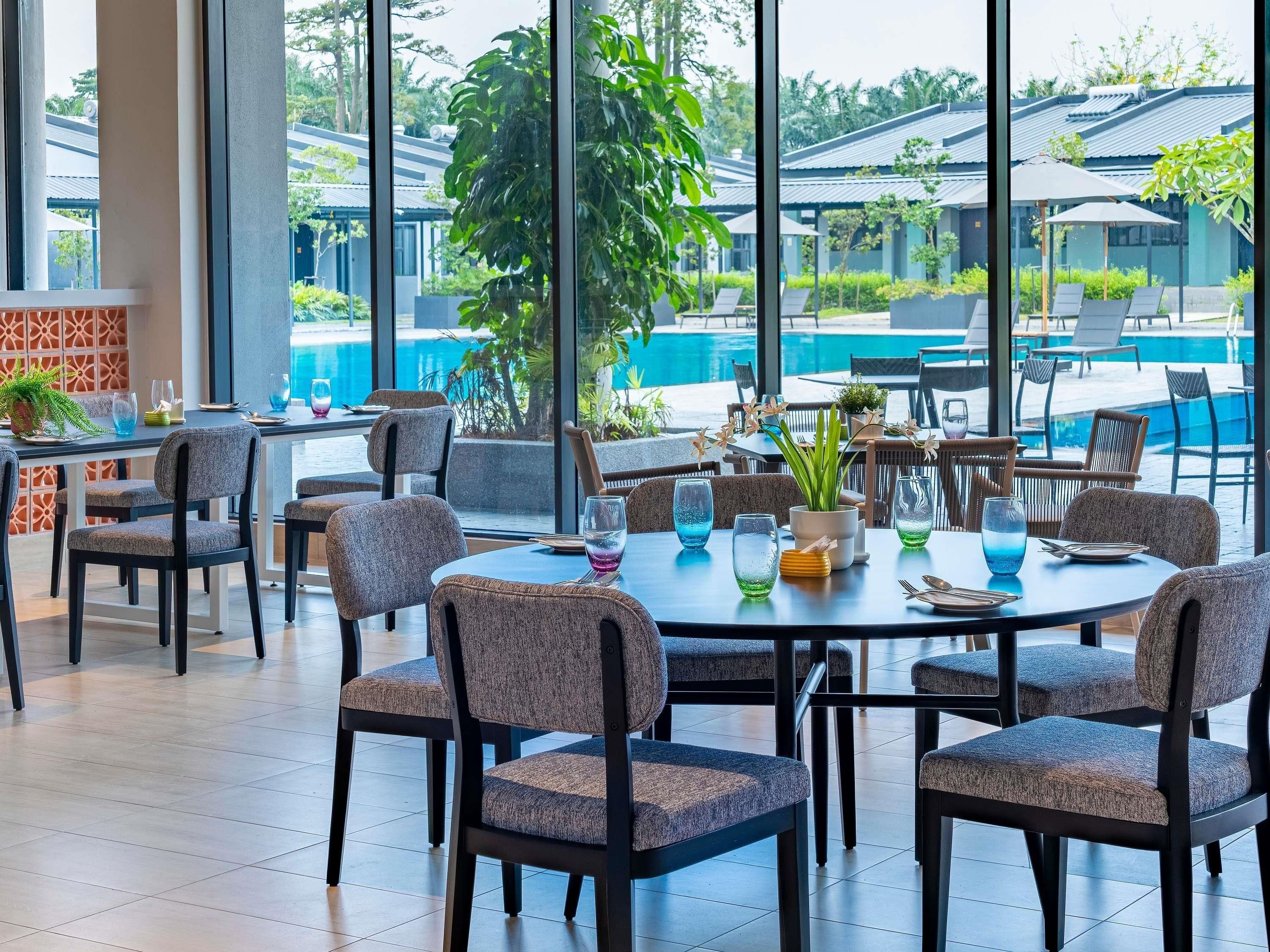 Um restaurante com mesas e cadeiras redondas, uma piscina ao fundo e grandes janelas.