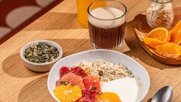 A breakfast setting with a bowl of yogurt topped with fruits and granola, a glass of orange juice, and a bowl of seeds.