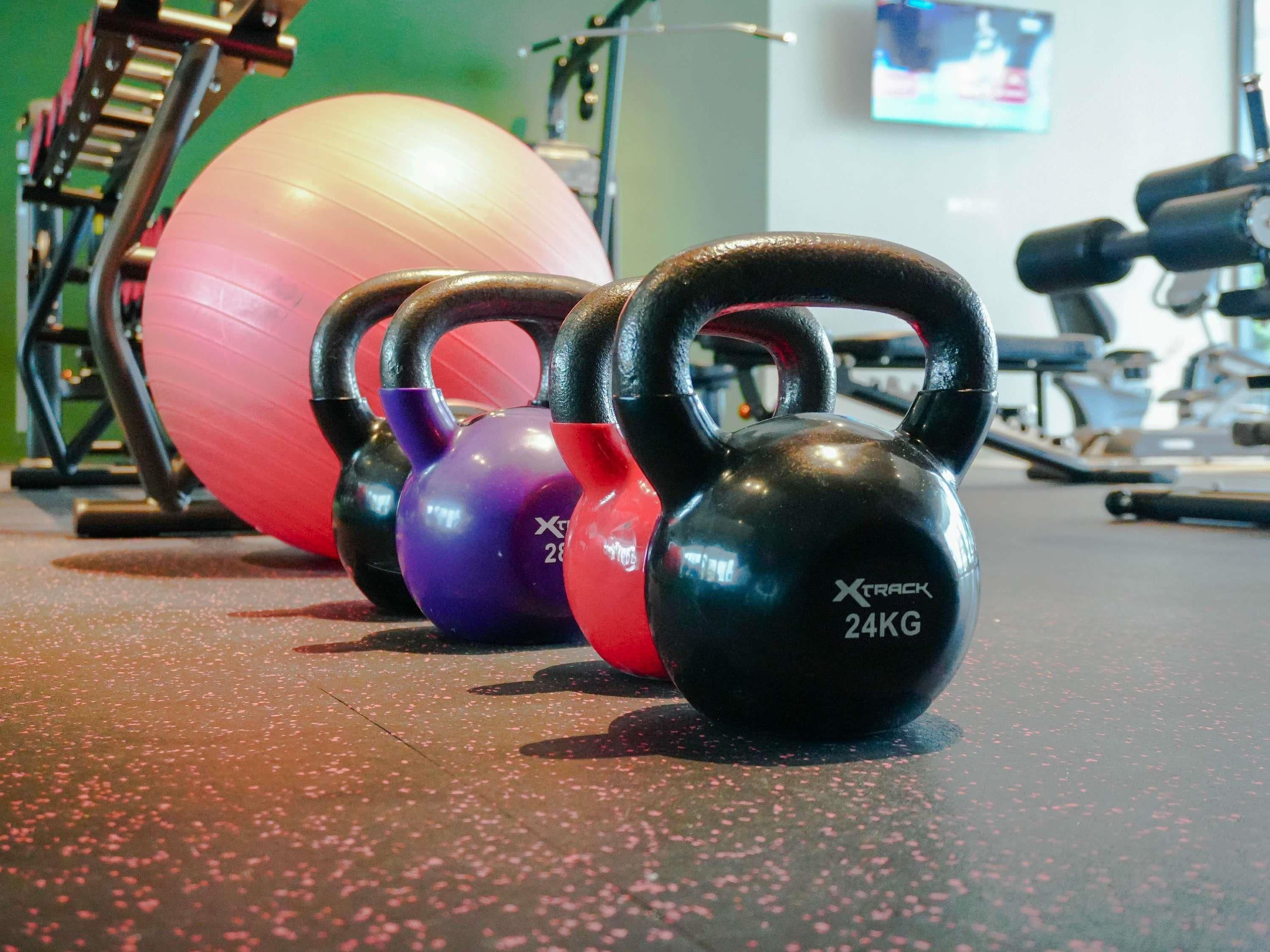 Varias mancuernas con asas y pesos, una pelota de ejercicios grande y equipo de gimnasio al fondo.