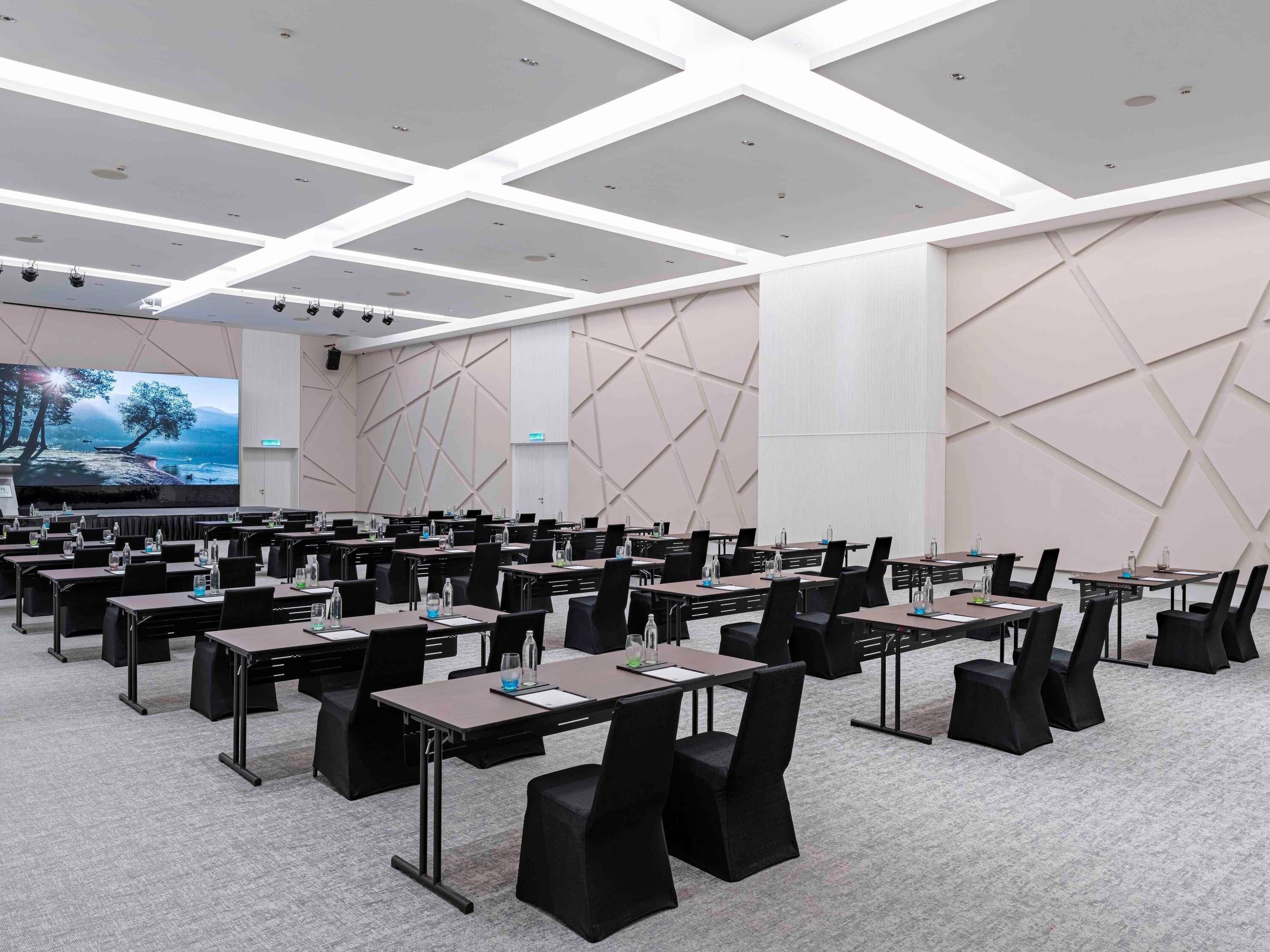 Een congreszaal met meerdere tafels en stoelen, een groot scherm en een wandontwerp met geometrische vormen.